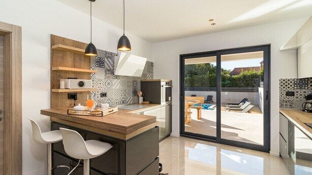 A modern kitchen with a wooden island, bar stools, and a large sliding glass door leading to an outdoor seating area.