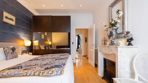 A modern bedroom with a large bed, bedside lamps, a mirror, a fireplace, and a white chair. - Charming flat Carré d'Or/Musiciens, 5 min away from the beach, WIFI (Nice)