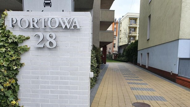 Ein Gebäudeeingang mit einem Schild, auf dem 'PORTOWA 28' steht, und einem gepflasterten Weg, der zum Eingang führt.