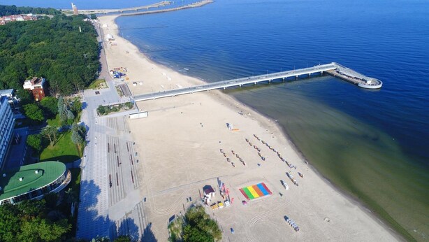 Eine Luftaufnahme eines Strandes mit einer langen Pier, die ins Wasser führt, einem sandigen Strandbereich mit Sonnenschirmen und einem Gebäude mit grüner Dachbegrünung auf der linken Seite.