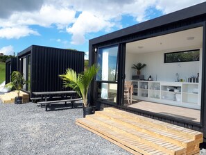 A modern house with a large sliding glass door leading to a kitchen area, a wooden deck, and a small outdoor seating area. - Aotearoa Surf Eco Pods (Te Arai)