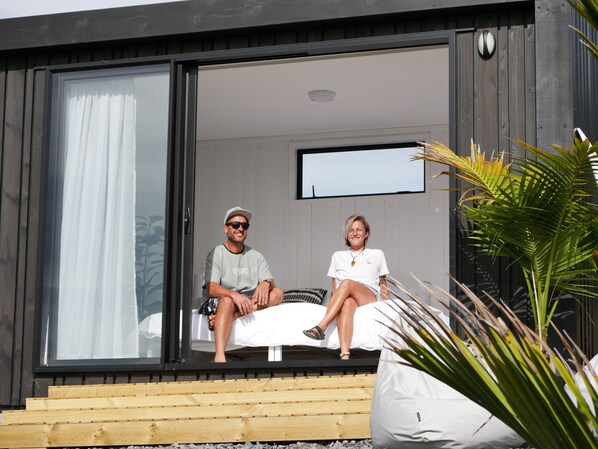 A modern outdoor seating area with a wooden deck and a glass door leading into a room. - Aotearoa Surf Eco Pods (Te Arai)