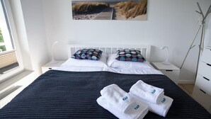 A neatly made bed with white linens and two towels with 'KAP' embroidered on them, a white headboard, and a large beach-themed painting above the bed. - Kap Ustronie Morskie - 365PAM (Ustronie Morskie)