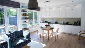 A modern kitchen with white cabinets, a dining area with a round table and chairs, and a large window overlooking a garden. - Kap Ustronie Morskie - 365PAM (Ustronie Morskie)