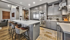 A modern kitchen with stainless steel appliances, a central island with bar stools, and a dining area with a gray sofa. - Exclusive Lakefront Luxury by Heavenly, Hot Tub | PEAK SS14 (South Lake Tahoe)
