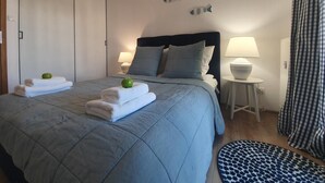 A neatly made bed with folded towels and a green apple, a bedside lamp, and a checkered curtain.