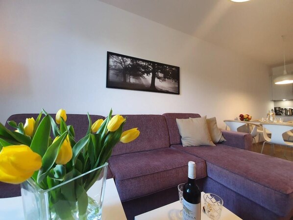 A living room with a purple sofa, a vase of yellow tulips, and a bottle of wine on a table. - Apartament Polanki Park - 365PAM (Kolobrzeg)