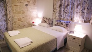 Una cama con una colcha texturizada, dos almohadas y dos toallas dobladas. Hay una mesita de noche con una lámpara y una cajita encima. La habitación tiene una pared de piedra y cortinas con estampado floral.