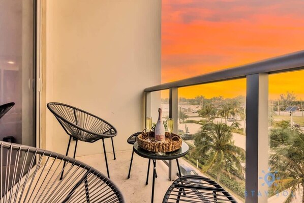 A balcony with two black wire chairs, a small round table with a bottle and glasses, and a view of palm trees and buildings.