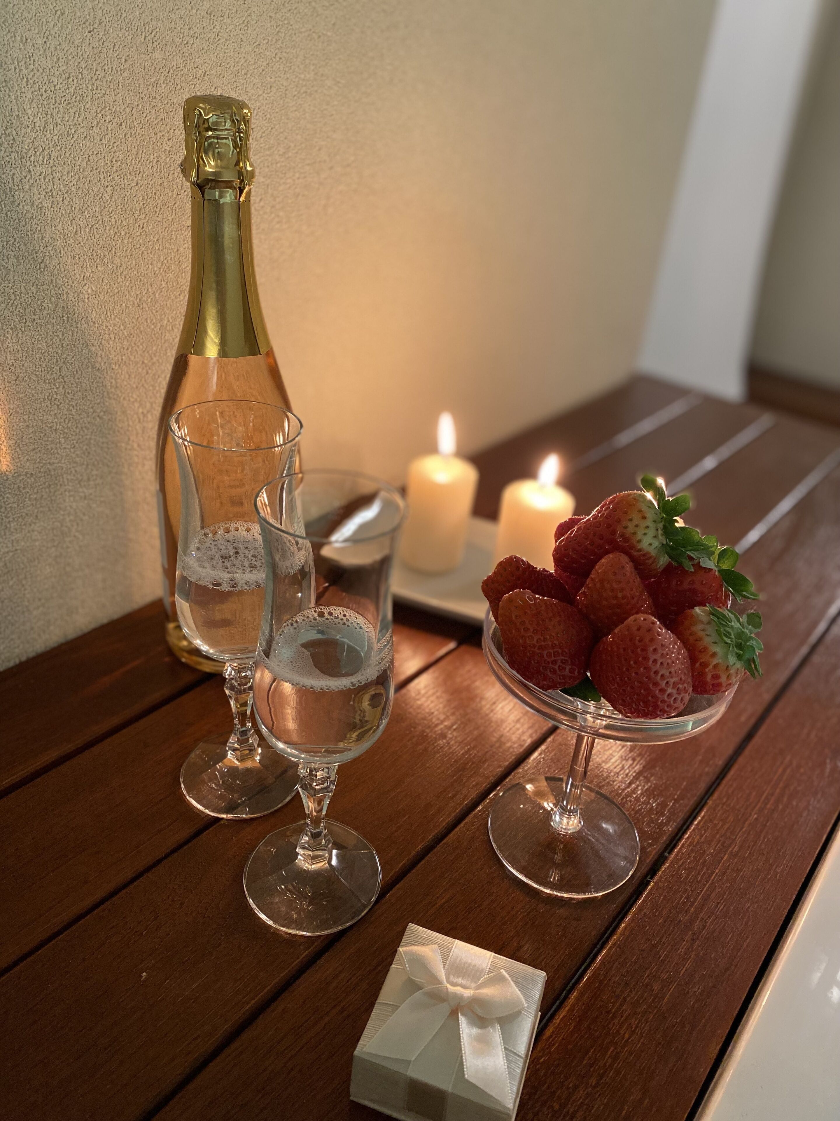 A wooden table with a bottle of champagne, two glasses, a candle, and a bowl of strawberries.
