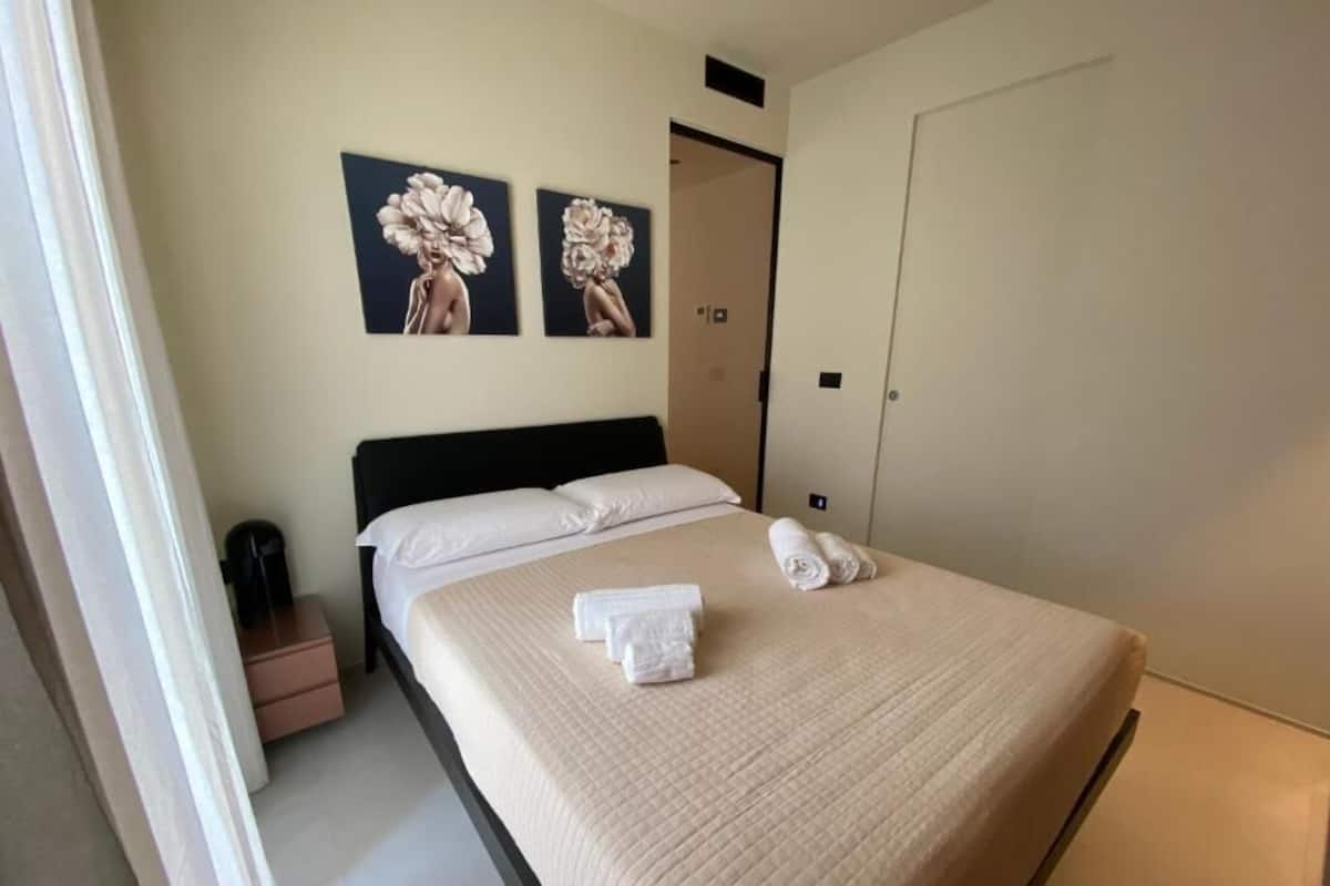 Una cama bien tendida con sábanas y toallas blancas, un cabecero negro y dos obras de arte con motivos florales en la pared.