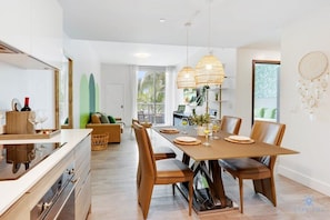 A modern dining area with a wooden table and leather chairs, a kitchen with white cabinets, and a large window with a view of palm trees.
