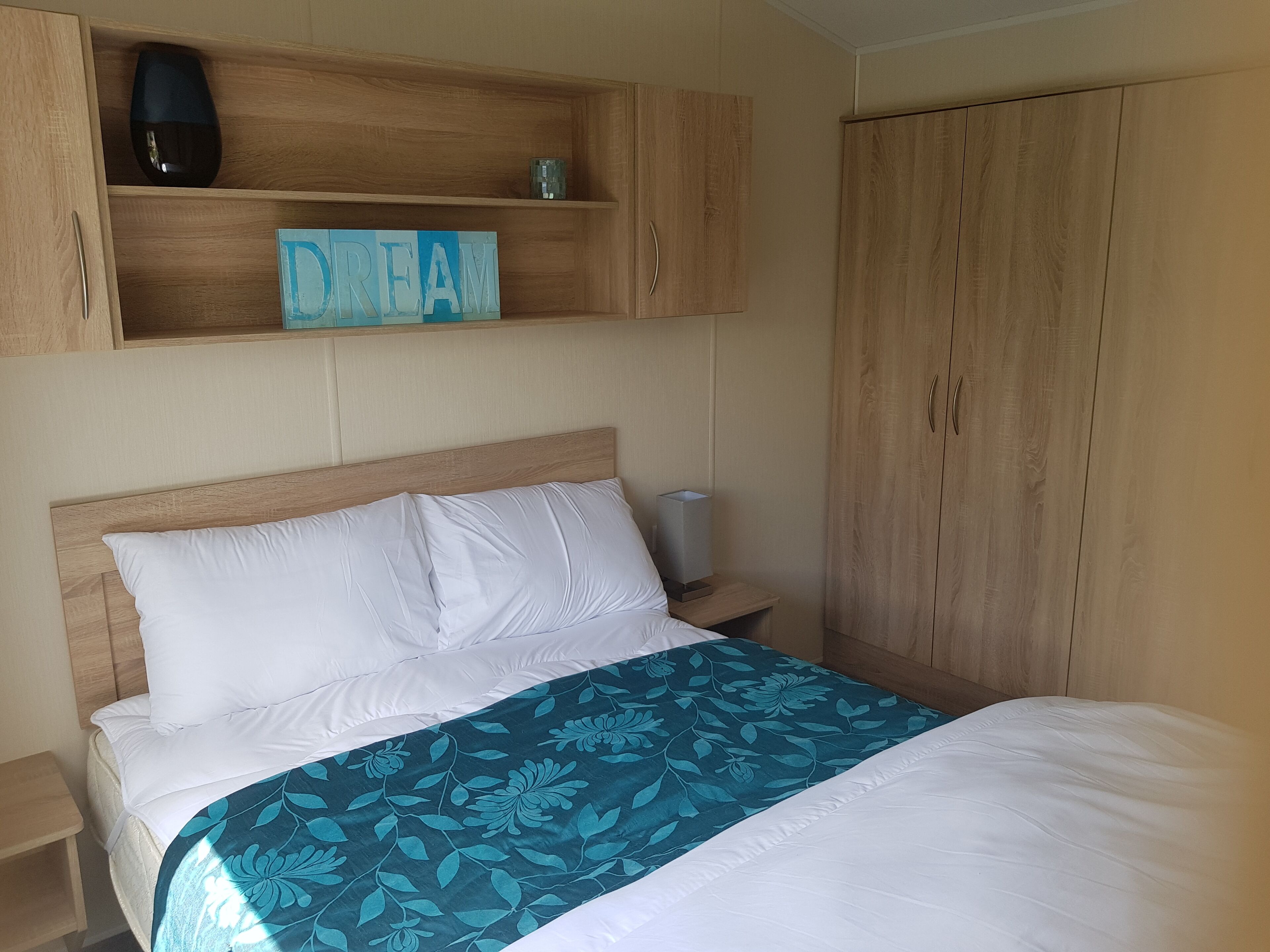 Een opgemaakt bed met een bloemenbedekker, een bedlampje en een plankje met de tekst 'DREAM'.