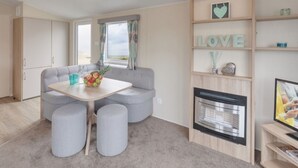 A modern living room with a sofa, ottomans, a table with fruit, a fireplace, and a TV. - Caravan - Sleeps6-pet Friendly-parking Available (Skegness)