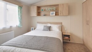 A bedroom with a bed, a nightstand, a shelf with a 'LOVE' sign, and a window with curtains. - Caravan - Sleeps6-pet Friendly-parking Available (Skegness)