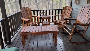 En träveranda med Adirondack-stolar och ett litet bord, med utsikt över ett skogsområde.