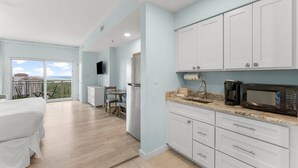 A modern kitchen with white cabinets, a microwave, and a coffee maker. There is a dining area with a table and chairs, and a balcony with a view. - Gulf Views ~ Studio ~ Resort Pool (Miramar Beach)