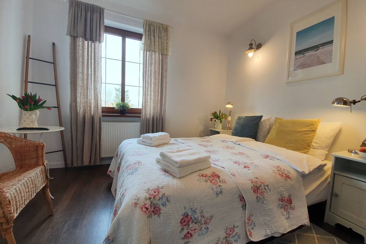 A neatly arranged bedroom with a bed, bedside tables, a chair, a window with curtains, a radiator, and a framed picture on the wall.
