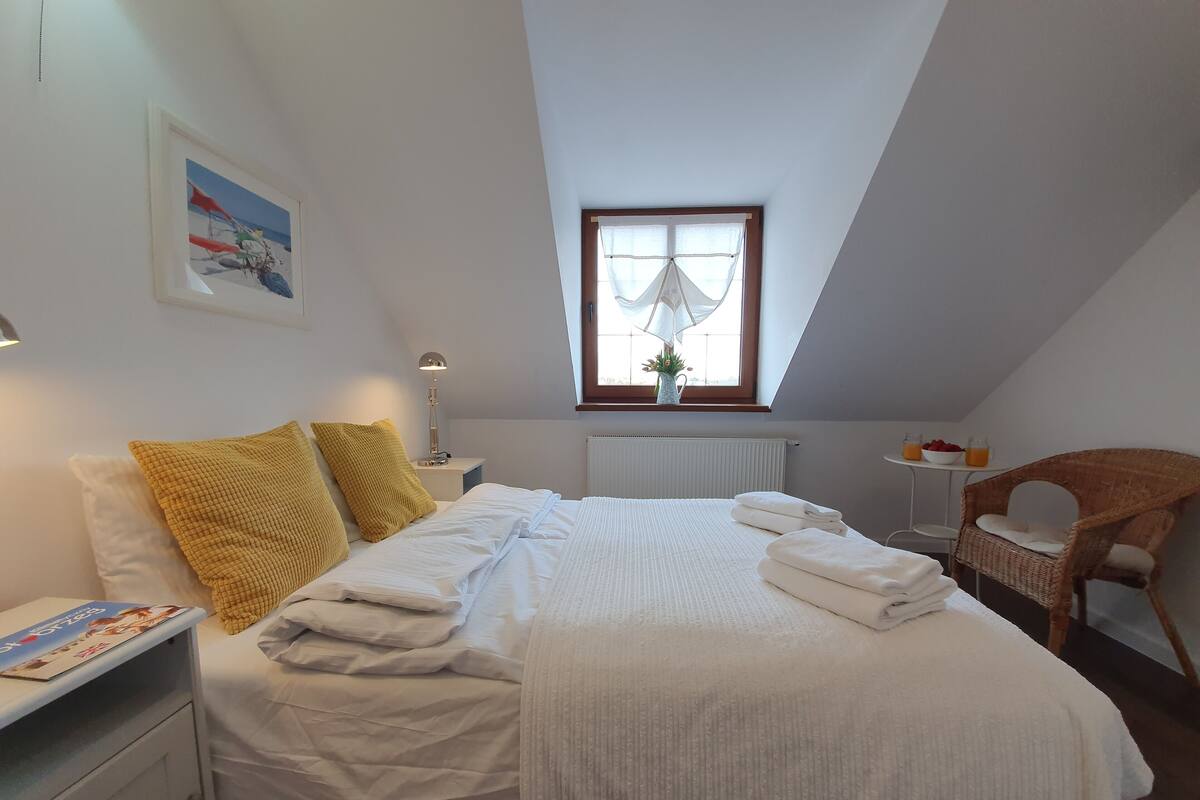A bedroom with a bed, pillows, a bedside table with a lamp, a window with curtains, a radiator, a chair, and a small table with a bowl and bottles.