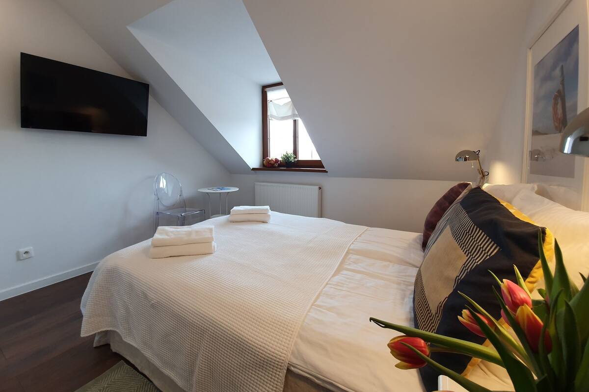 A bedroom with a large bed, a TV mounted on the wall, a small table, and a window with a view of the sky.