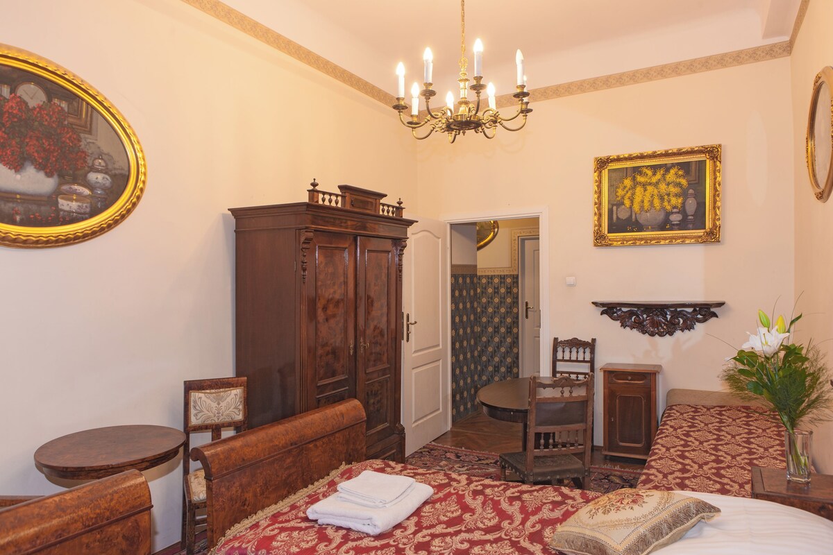 A bedroom with a wooden wardrobe, a bed with a red and white patterned bedspread, a wooden chair, a small round table, and a chandelier.