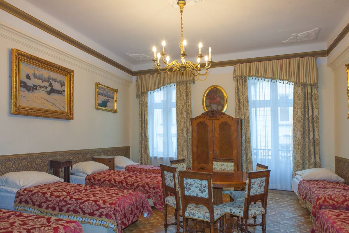 A hotel room with four beds, a wooden dining table, and chairs. There is a chandelier, a large mirror, and two windows with curtains.