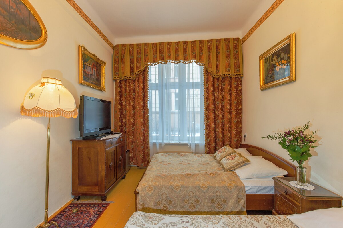 Habitación de hotel con cama, televisión, mesita de noche, lámpara y ventana con cortinas.