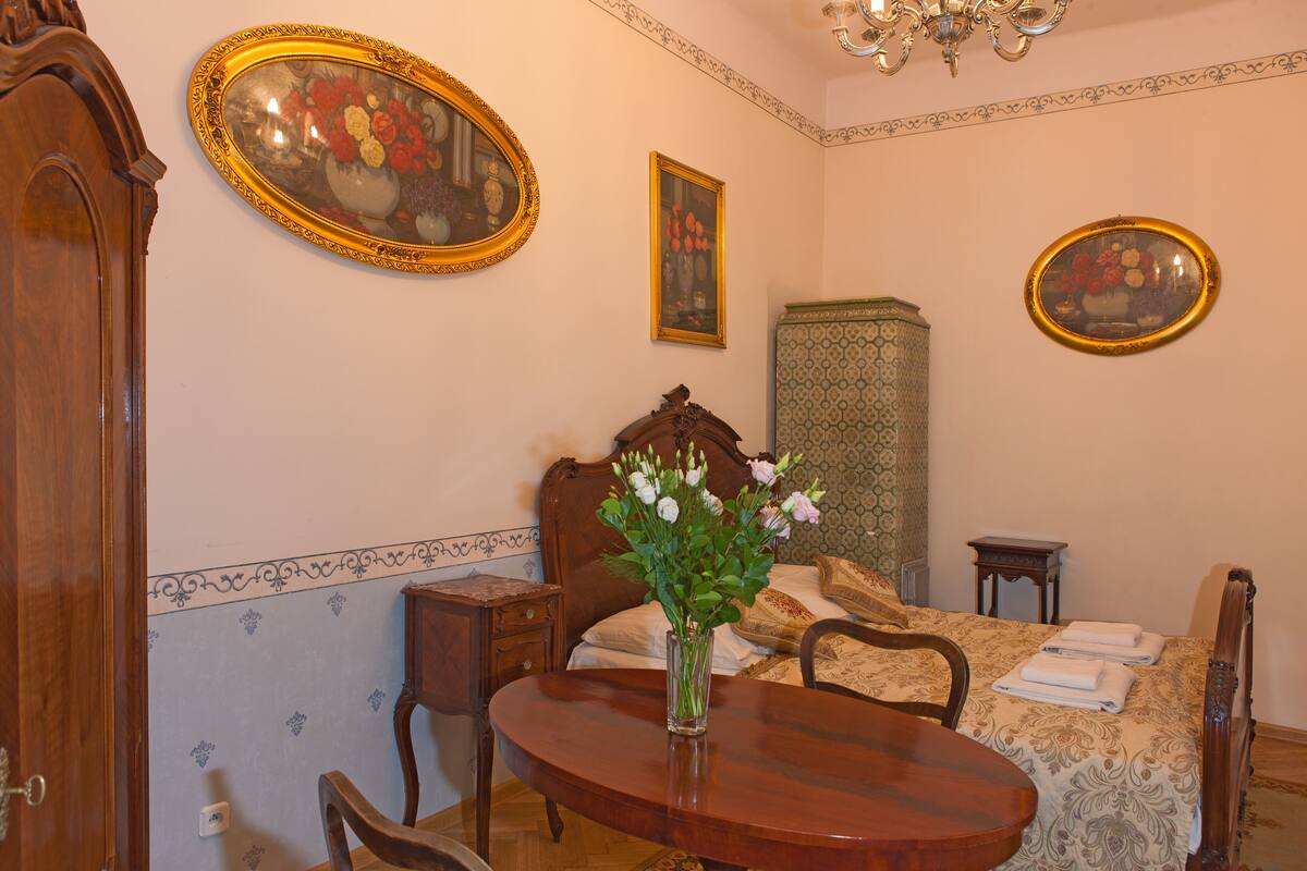 A bedroom with a bed, a bedside table, a chair, a wooden table with a vase of flowers, and a mirror on the wall.
