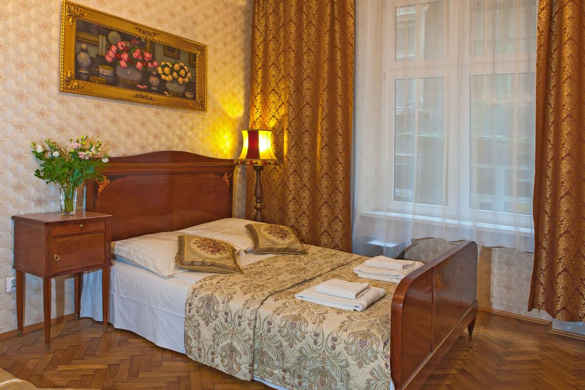 A bedroom with a wooden bed, a nightstand, a lamp, a vase with flowers, and a large window with curtains.