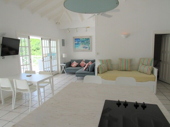 A spacious living area with a dining table, white chairs, a sofa with colorful cushions, and a television mounted on the wall.