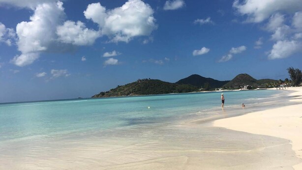 A beach with clear turquoise water, white sand, and distant green hills under a partly cloudy sky. - Jolly Harbour, 3 bedroom villa, pool, close to beach and all amenities. (Jolly Harbour)