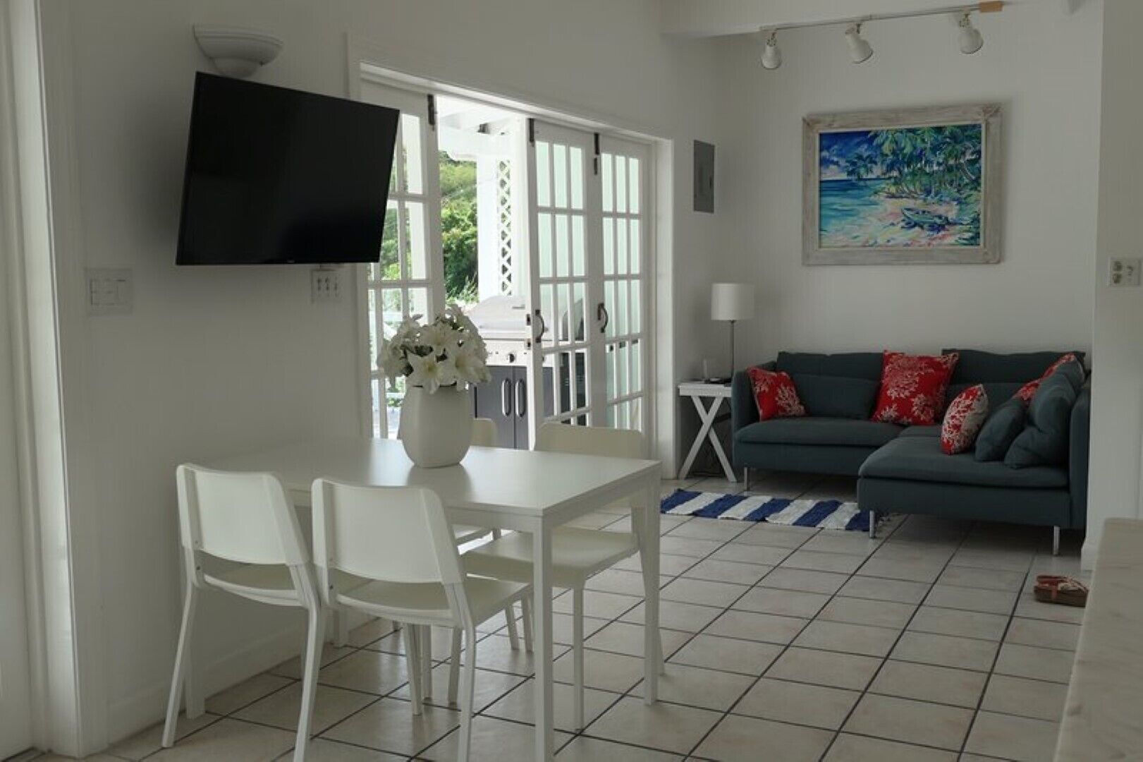 A living room with a white dining table, two white chairs, a grey sofa with red and grey cushions, a TV mounted on the wall, and a painting of a beach scene.