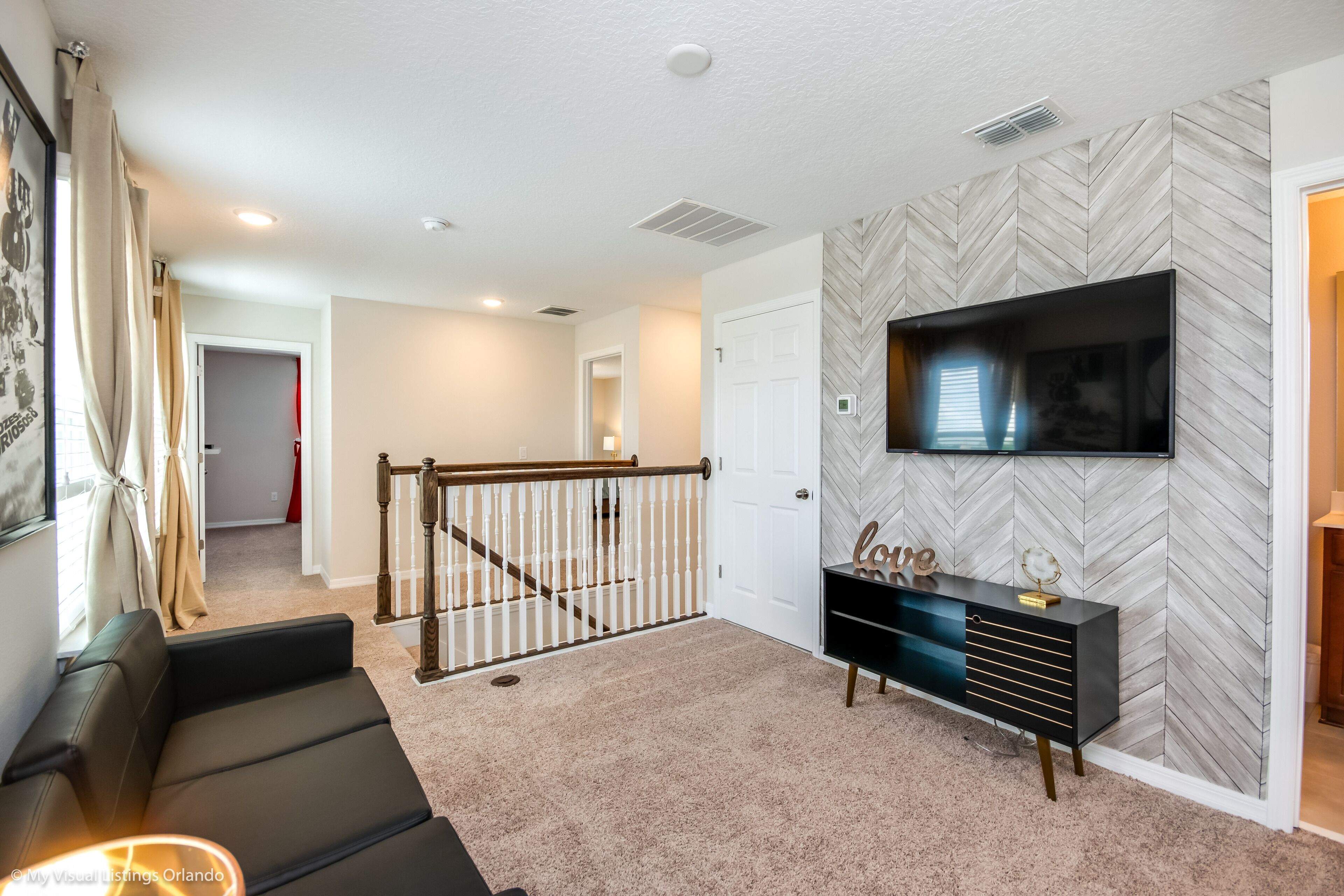 A modern living room with a herringbone-patterned wall, a flat-screen TV mounted above a black console, a staircase leading to another room, and a beige carpet.
