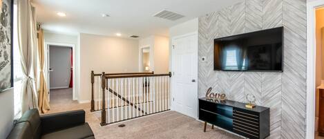 A modern living room with a herringbone-patterned wall, a flat-screen TV mounted above a black console, a staircase leading to another room, and a beige carpet.