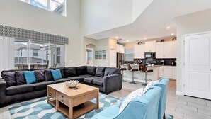 A spacious living area with a sectional sofa, a coffee table, and a blue armchair. A kitchen is visible in the background with white cabinets and a microwave.