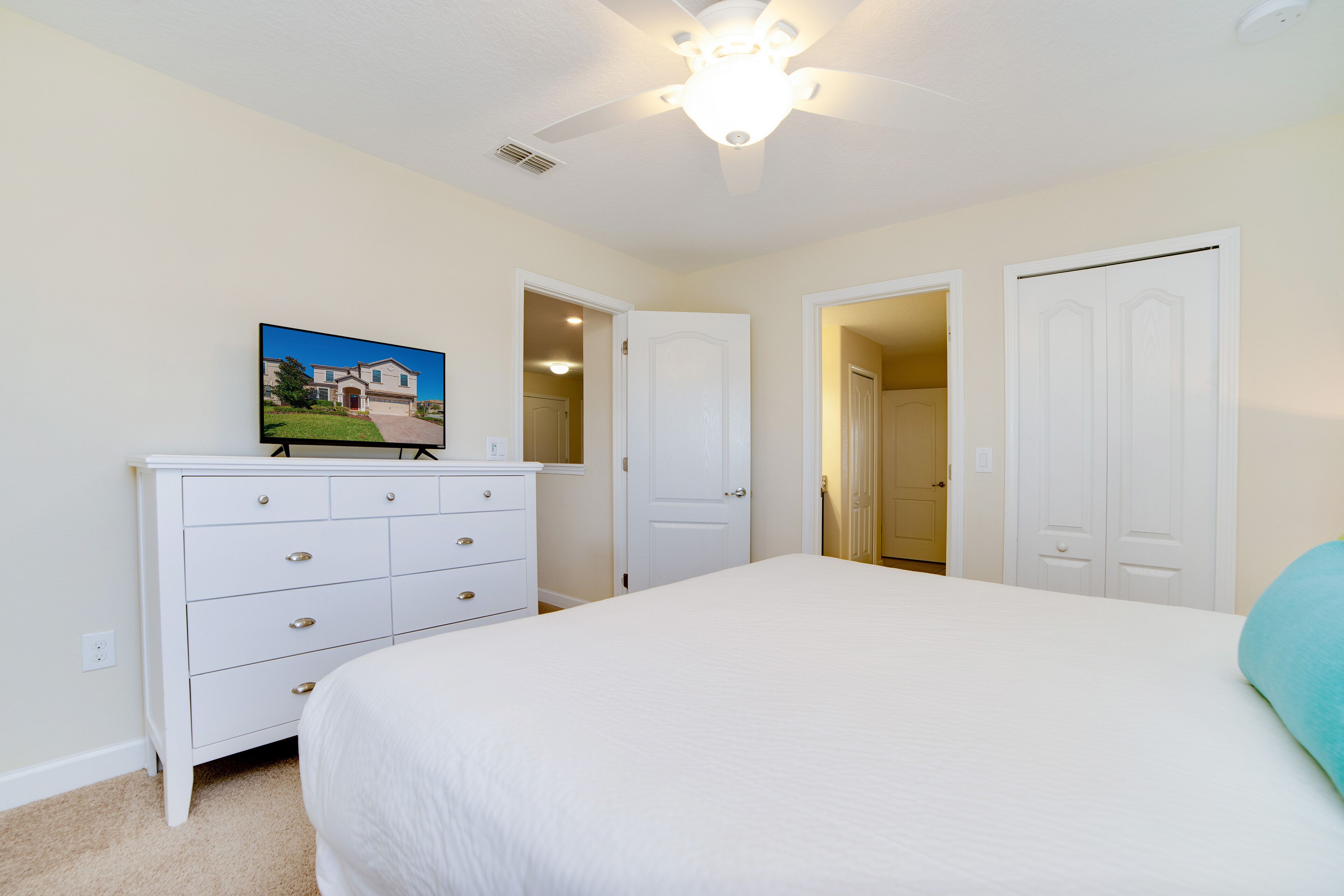 A bedroom with a bed, a dresser, a ceiling fan, and a door leading to another room.