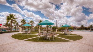 Un espace extérieur aménagé en terrasse, avec des tables et des chaises sous des parasols, entouré de palmiers et agrémenté d’une fontaine.