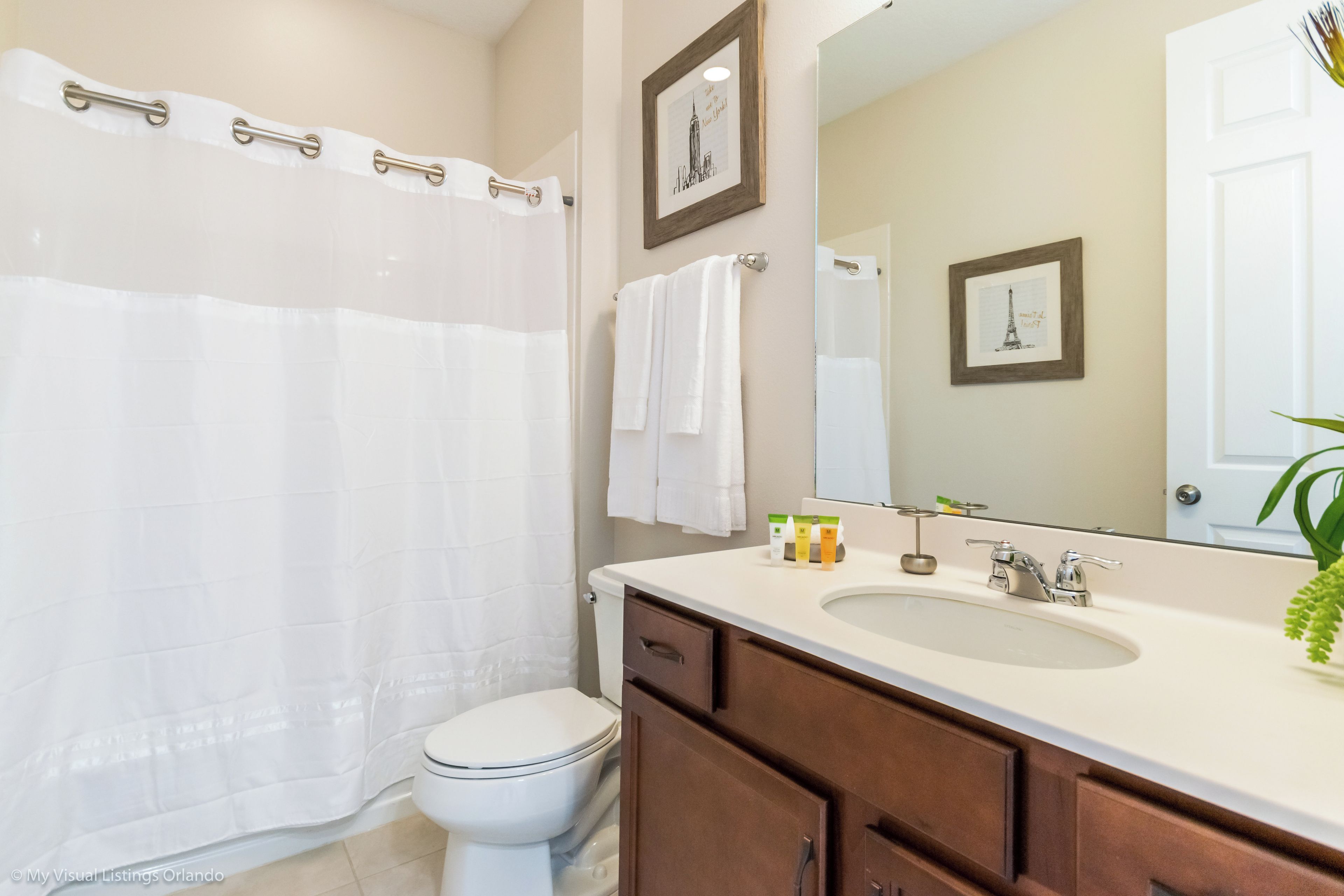 A bathroom with a shower curtain, toilet, sink, vanity, and two framed pictures on the wall.