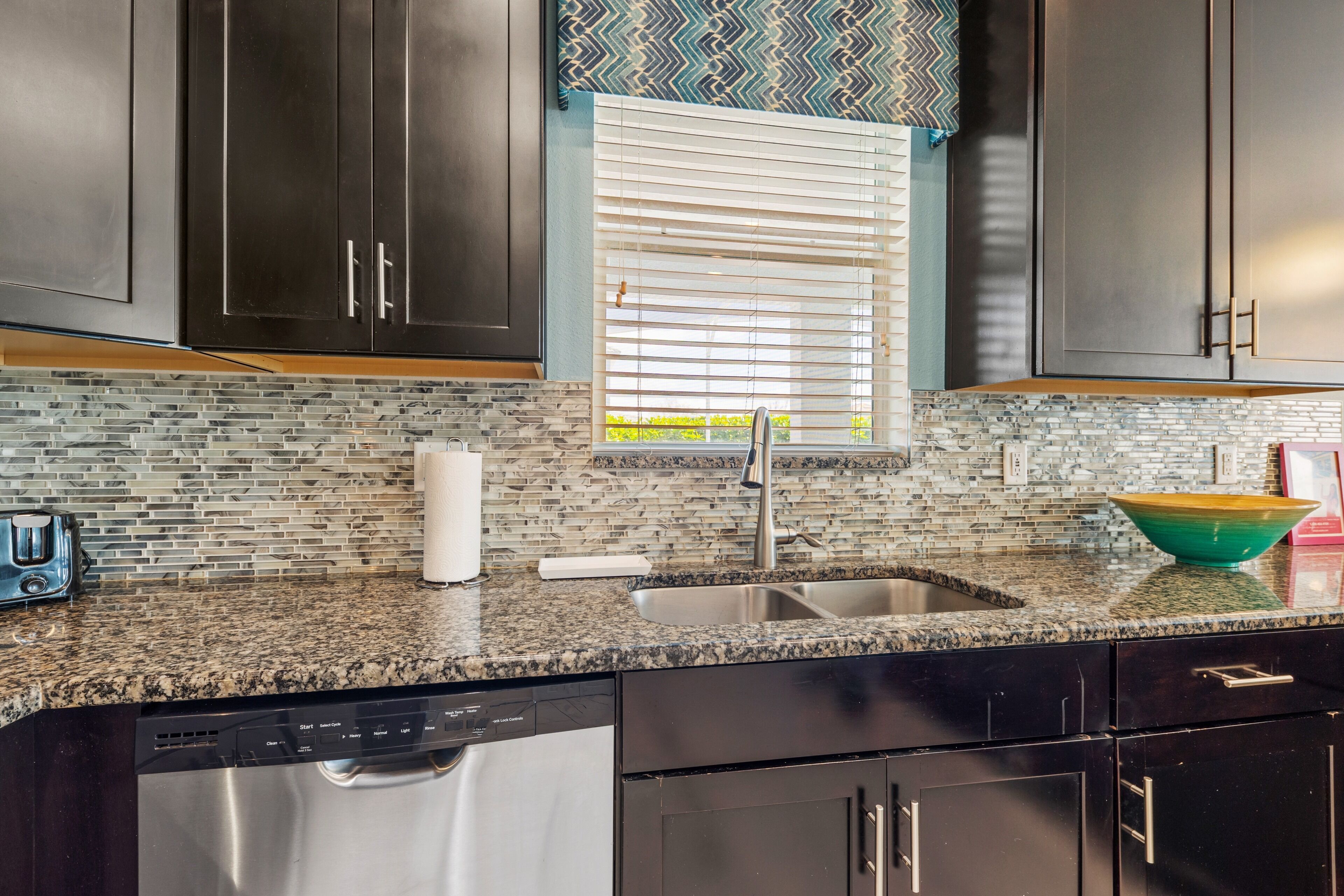 A kitchen with dark cabinets, a granite countertop, a stainless steel sink, and a window with blinds.