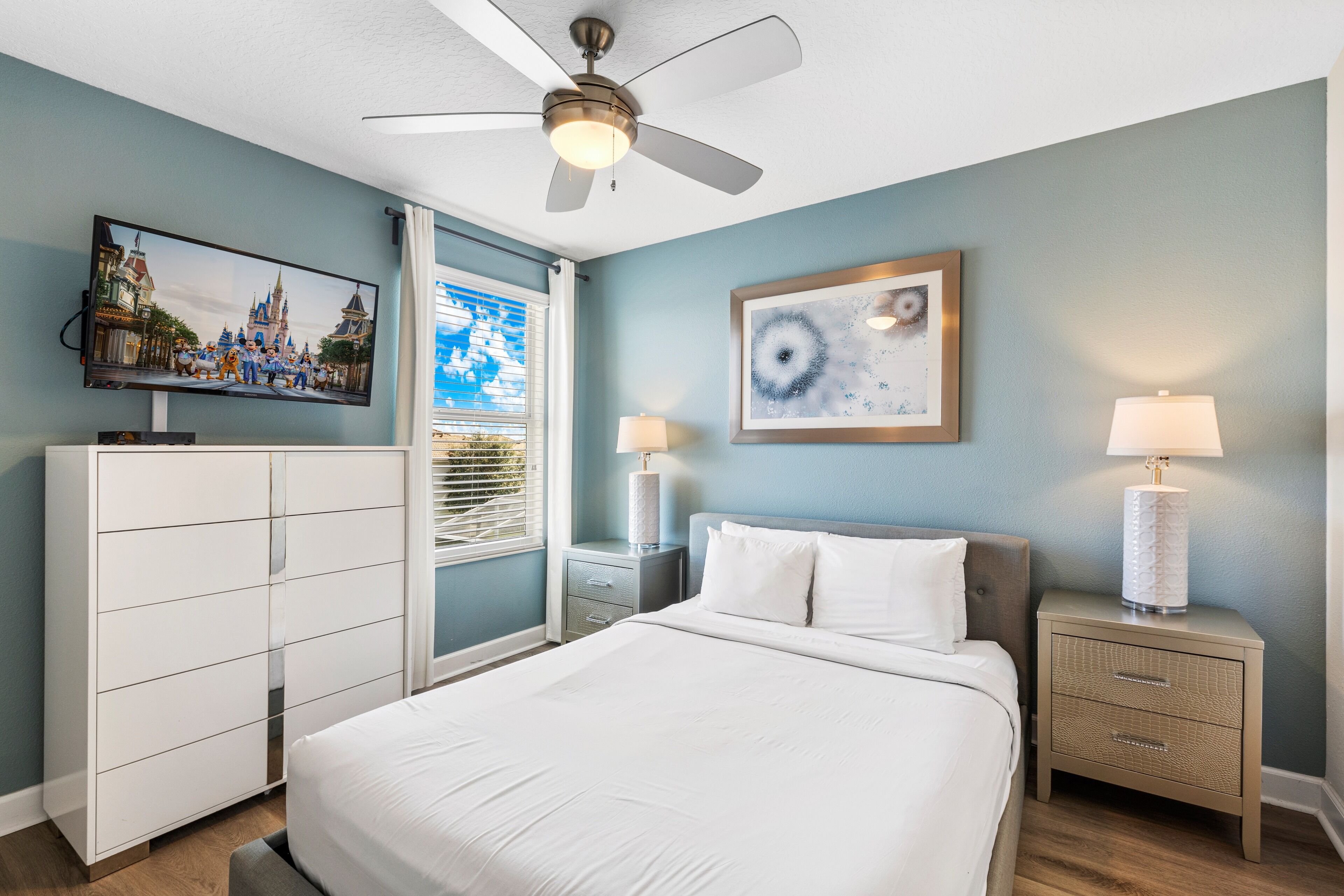 A bedroom with a bed, a ceiling fan, a TV on the wall, a nightstand with a lamp, and a window with blinds.