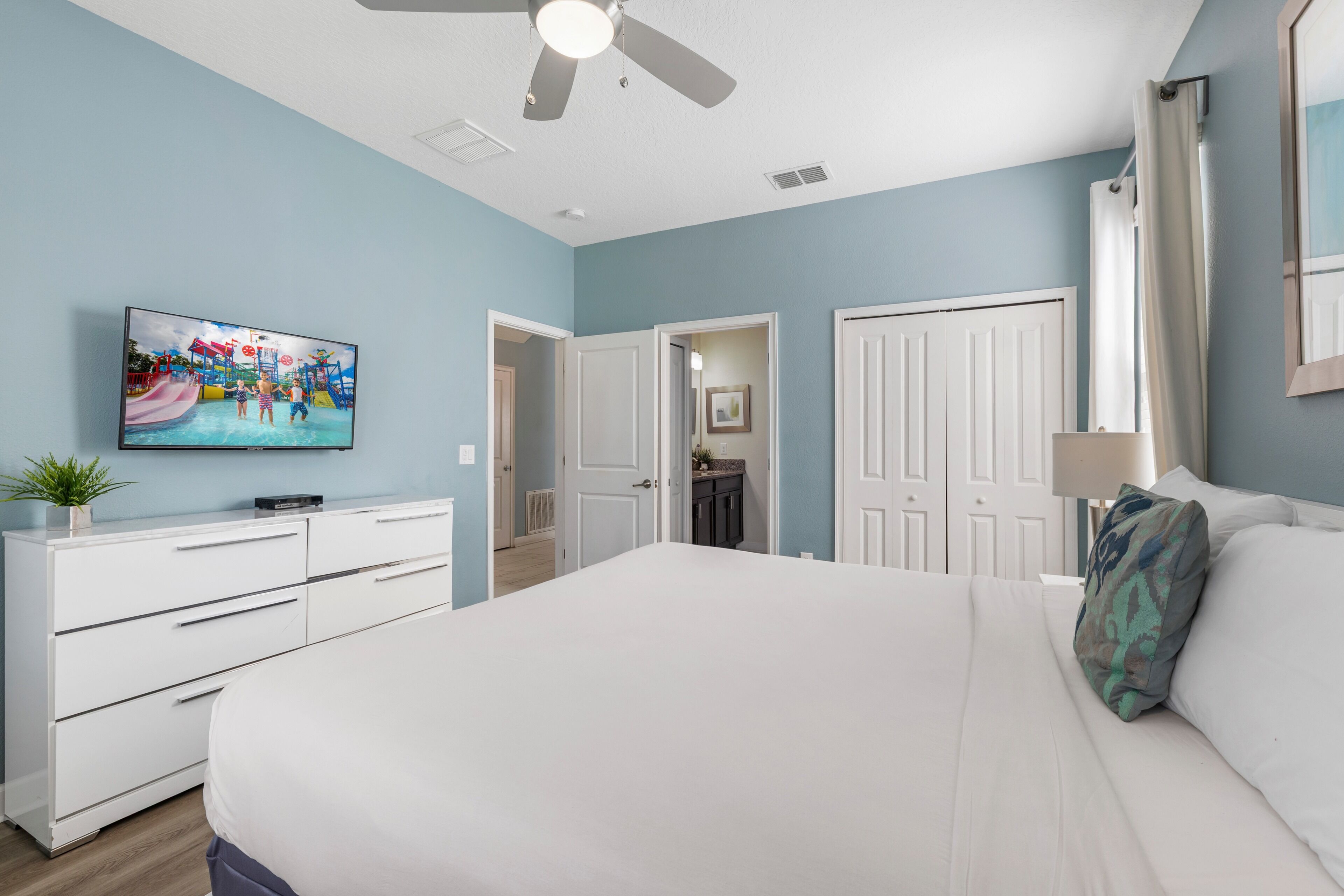 A bedroom with a large bed, a ceiling fan, a TV on the wall, and a door leading to another room.