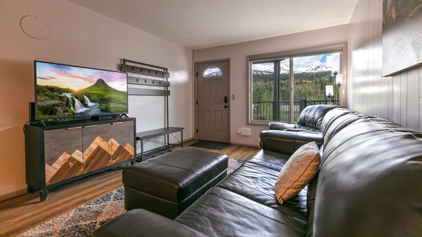 A living room with a large flat-screen TV, a black leather sofa, and a wooden cabinet with a mountain landscape picture. - Gold Creek Condo: Downtown, Amazing Views (Breckenridge)