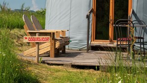 En träveranda med en skylt som visar texten ”COQUÉLICOT”, som leder till ett takförsett utomhusområde med en trästol och ett bord.