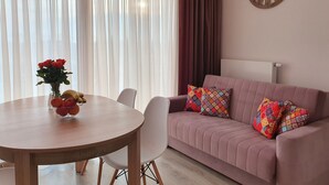 Una sala de estar con una mesa redonda de madera, sillas blancas, un sofá rosa con almohadones coloridos, un reloj de pared y una ventana con cortinas translúcidas.