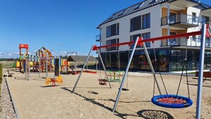 Un parque infantil con columpios y un neumático colgante, un edificio con balcones y una bandera.