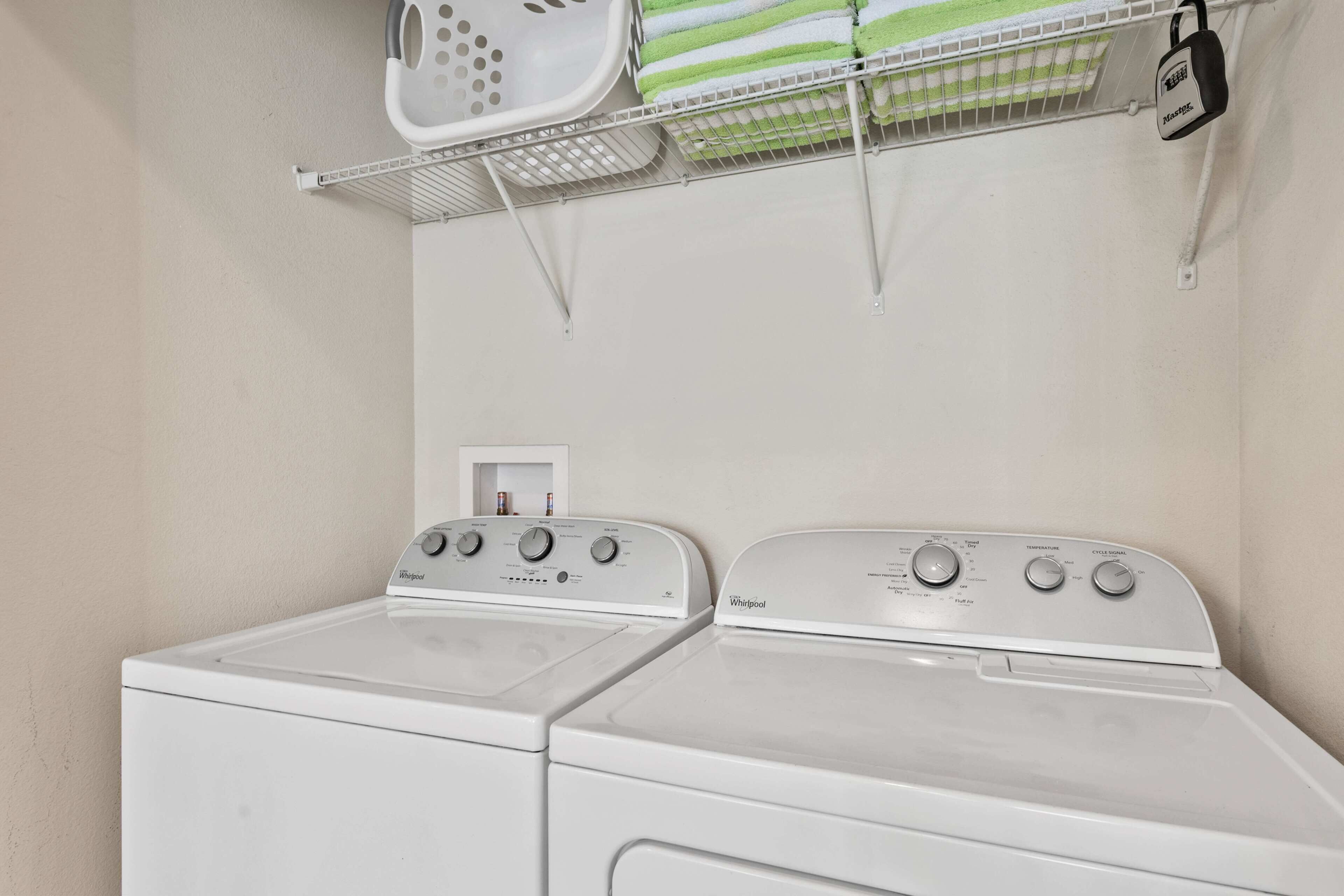 A laundry area with two white Whirlpool washers and a metal shelf above holding laundry baskets and a white appliance.
