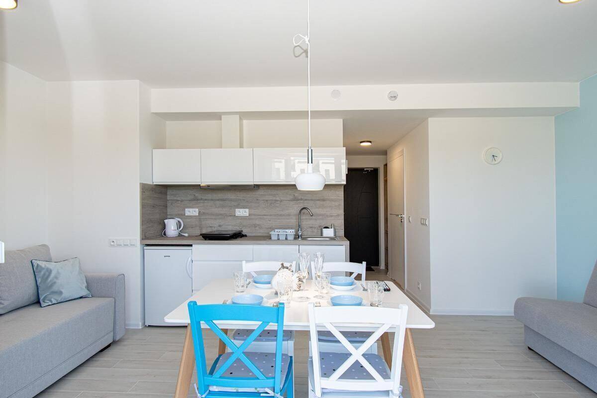 Une cuisine moderne avec des meubles blancs, un plan de travail blanc et un four encastré. On y trouve une table à manger avec des chaises bleues et une nappe blanche.