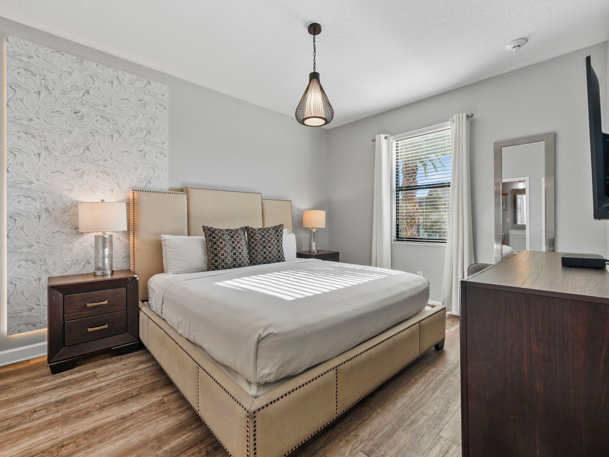 A bedroom with a large bed, two bedside tables with lamps, a dresser, and a window with blinds.