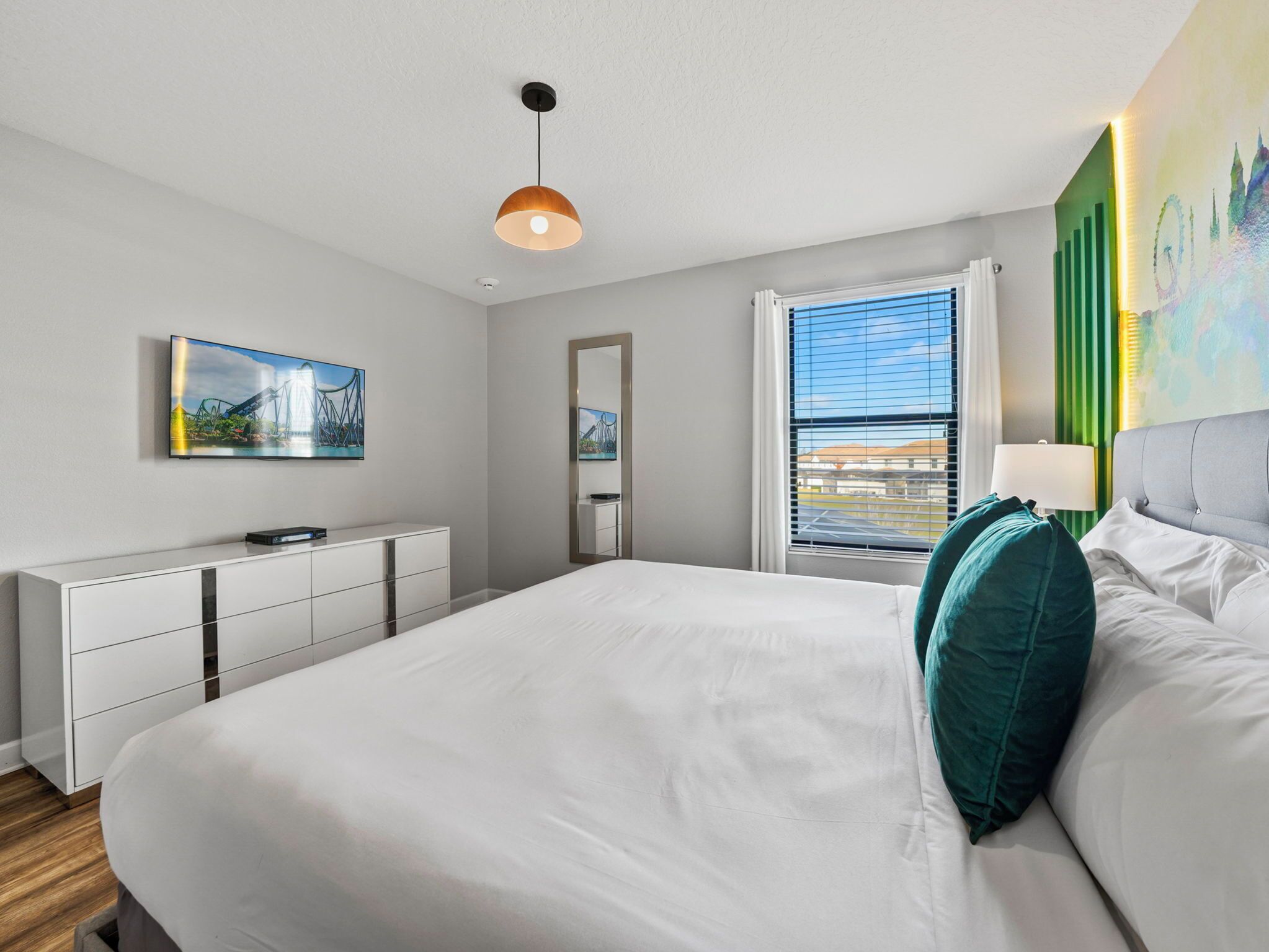 A modern bedroom with a large bed, a dresser, a television, and a window with blinds.