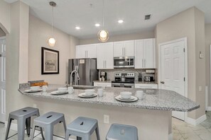 A modern kitchen with white cabinets, granite countertops, and stainless steel appliances. There are bar stools and a framed picture on the wall. - Resort Oasis! 3BR w Pool Access near Disney World (Kissimmee)